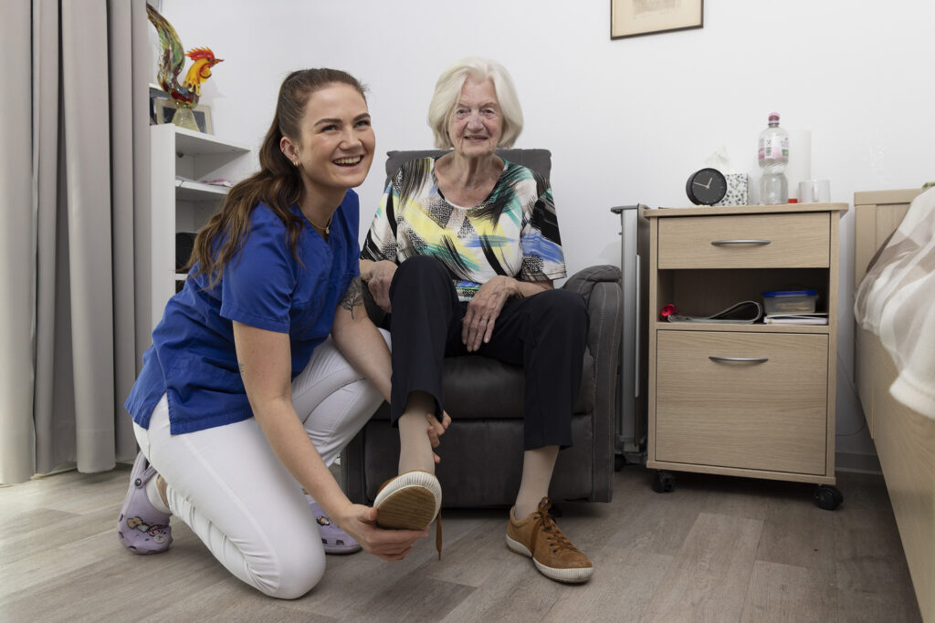 Eine Mitarbeiterin hilft einer Bewohnerin die Schuhe anzuziehen.