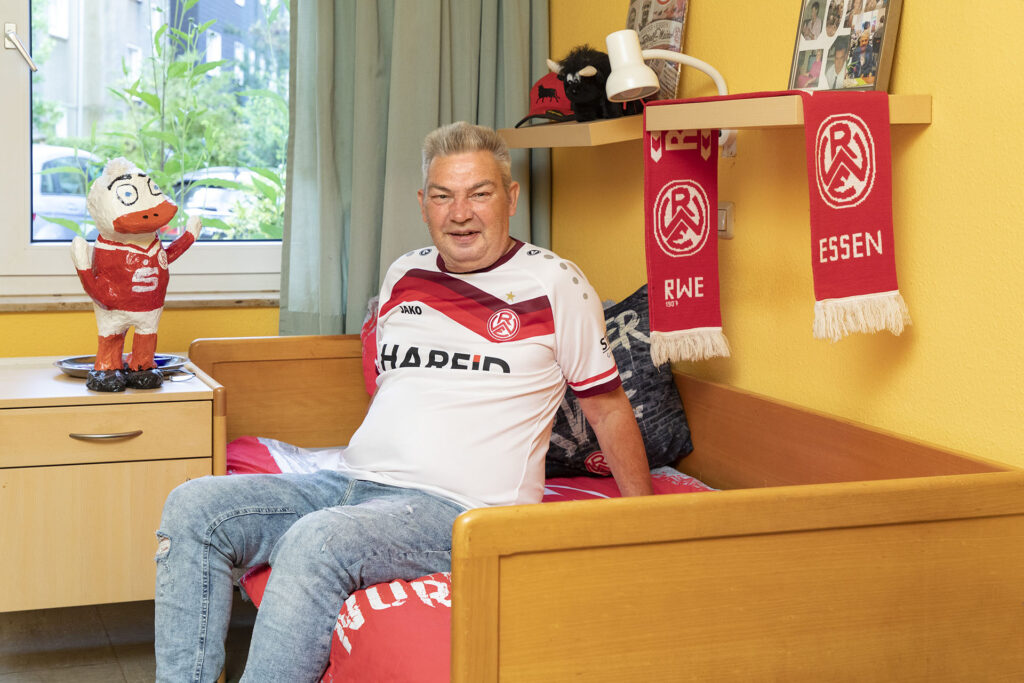 Ein Bewohner im Fußballtrikot zeigt sein Zimmer, das mit vielen Dingen vom Fußballverein Rot Weiss Essen dekoriert ist.