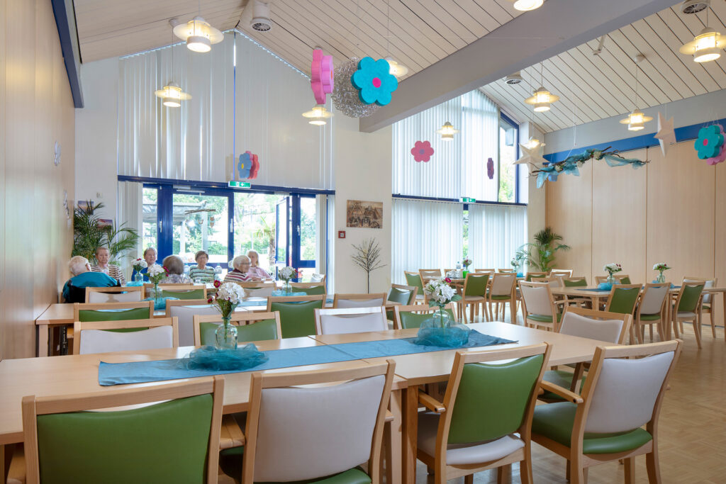 Blick in einen Teil des großen Saals, der mit zahlreichen Tischgruppen Platz für viele Bewohner und ihre Gäste bietet. Bodentiefe Fenster sorgen für Helligkeit und freundliche Dekoration für Gemütlichkeit.
