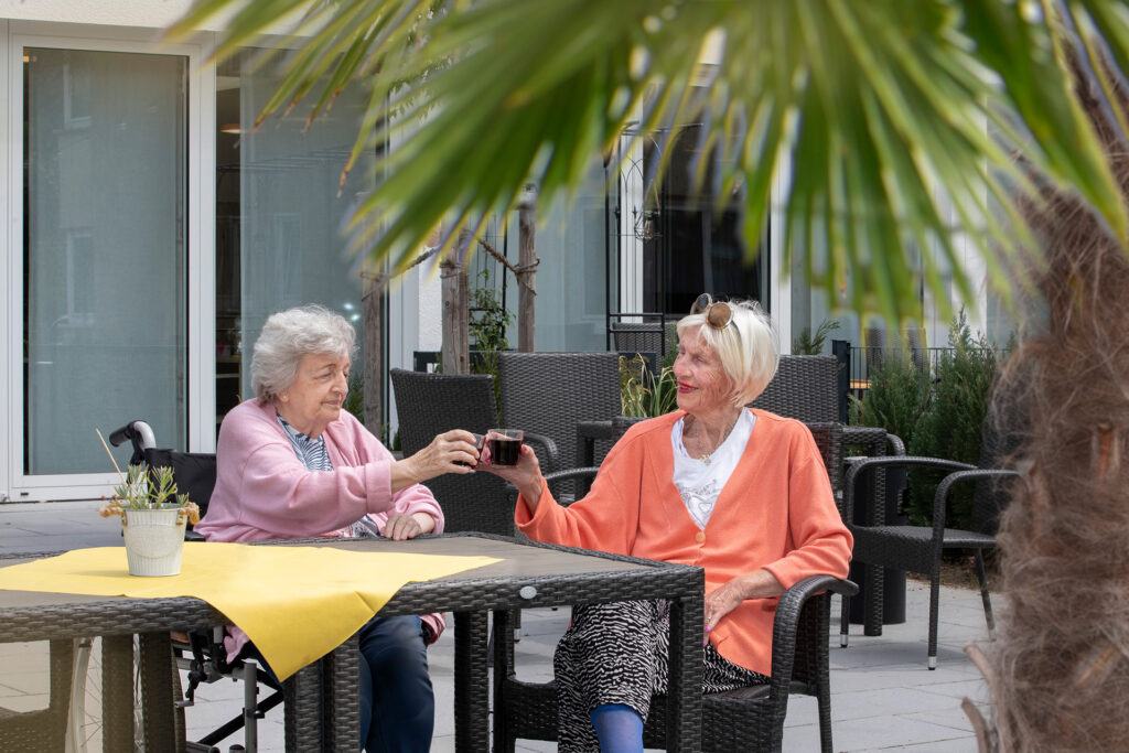Zwei Bewohnerinnen stoßen auf der Sonnen-Terrasse mit Getränken an.