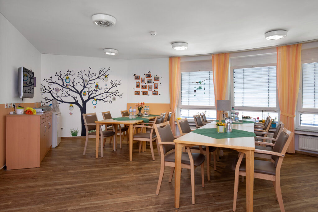 Blick in einen Aufenthaltsraum, in dem zwei Tischgruppen stehen. An der Wand sind hübsche Dekorationen, Fotos und ein Fernseher angebracht.