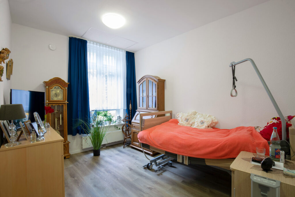 Blick in ein Bewohnerzimmer: Neben einem Bett ist genug Platz für die individuellen, antiken Möbel der Bewohnerin. Zahlreiche persönliche Gegenstände sorgen für Gemütlichkeit. Durch das große Fenster fällt viel Tageslicht in das Zimmer.