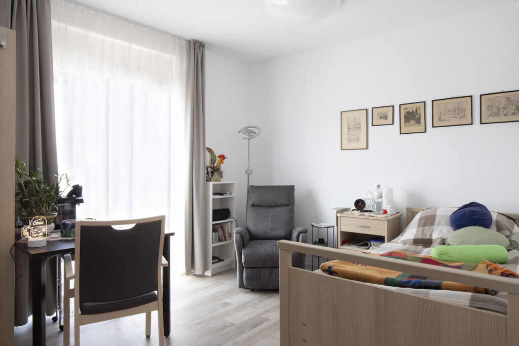 Blick in ein Zimmer: Bodentiefe Fenster lassen viel Tageslicht ins Zimmer. Neben einem Bett ist genügend Platz für weiteres individuelles Mobiliar und persönliche Gegenstände.