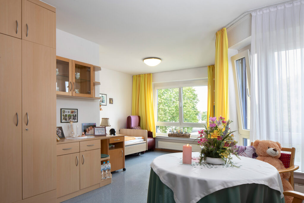 Blick in ein Zimmer: Große Fenster lassen viel Tageslicht in den Raum. Neben Schränken, einem Bett, Tisch und einem Fernsehsessel ist noch viel Platz im Zimmer.