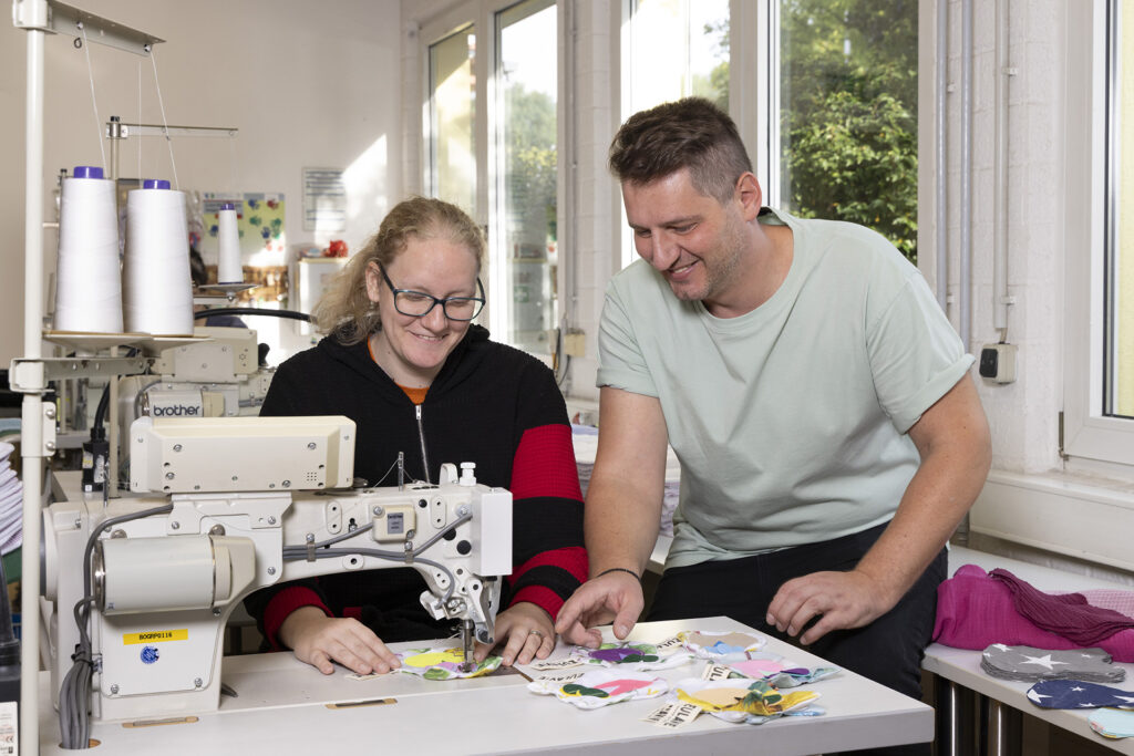 Eine Werkstattmitarbeiterin und ein Gruppenleider besprechen an der Nähmaschine die nächsten Schritte beim Nähen.