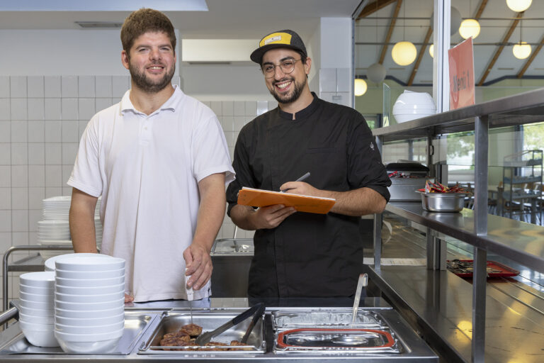 Zwei Werkstattmitarbeitende kontrollieren in der Küche mit einem Thermometer die Temperatur der Mahlzeiten.
