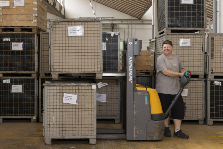Ein Werkstattmitarbeiter bewegt im Lager mit einem Palettenhubwagen große Gitterboxen.