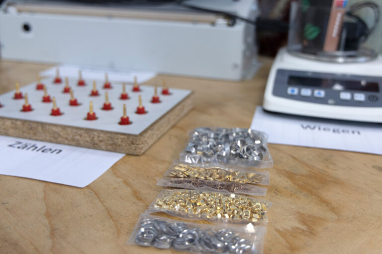 Verschiedene Muttern und Unterlegschrauben sind zu sehen. Im Hintergrund liegen zwei Zettel, auf denen "Zählen" und "Wiegen" steht. Dahinter steht eine Waage.