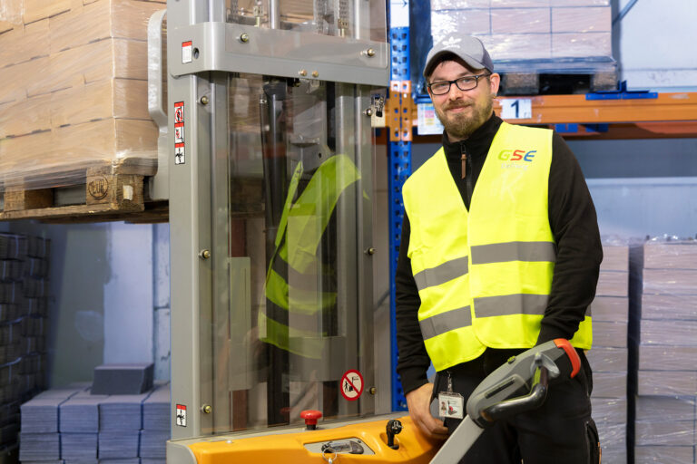 Ein Werkstattmitarbeiter arbeitet mit einem Hubwagen im Lagerbereich. Er trägt eine neongelbe Sicherheitsweste mit GSE-Logo darauf.