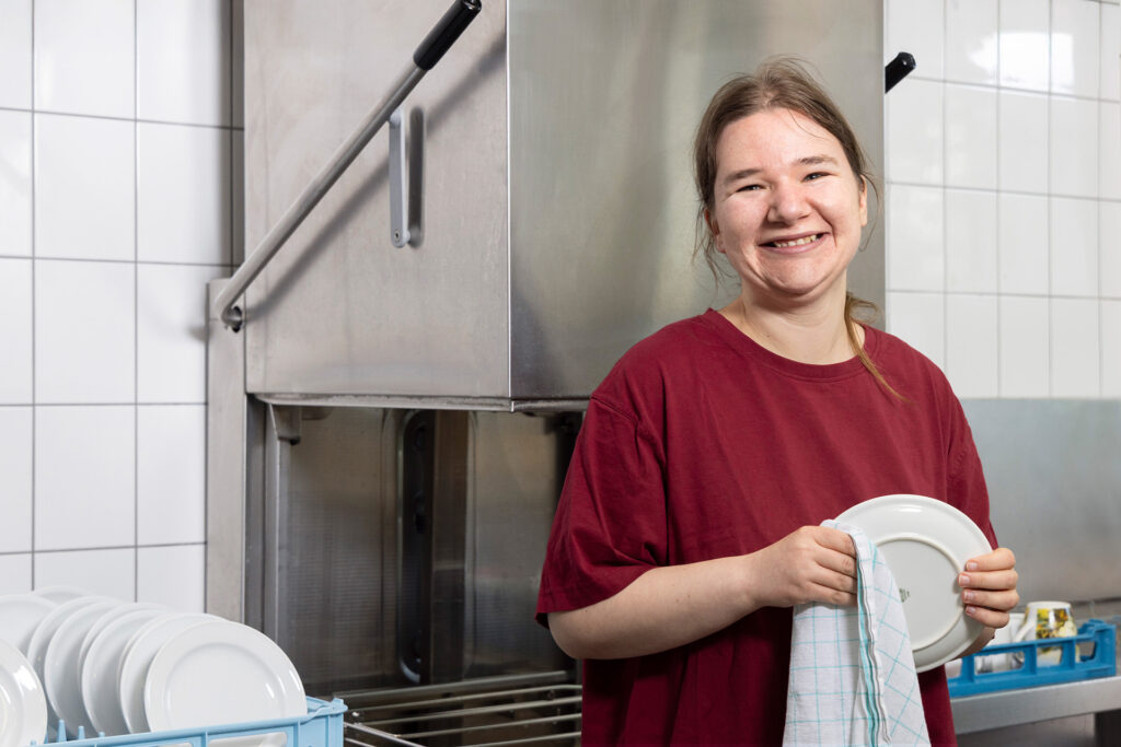 Eine junge Frau trocknet in einer Küche Geschirr ab und lächelt freundlich in die die Kamera.