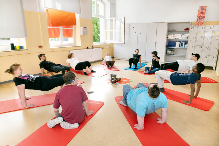 Morgens können die Teilnehmer am Yoga teilnehmen. Zu sehen sind mehrere Personen, die ihre Yogamatten in einem Kreis ausgelegt haben und hier gemeinsam Übungen machen.