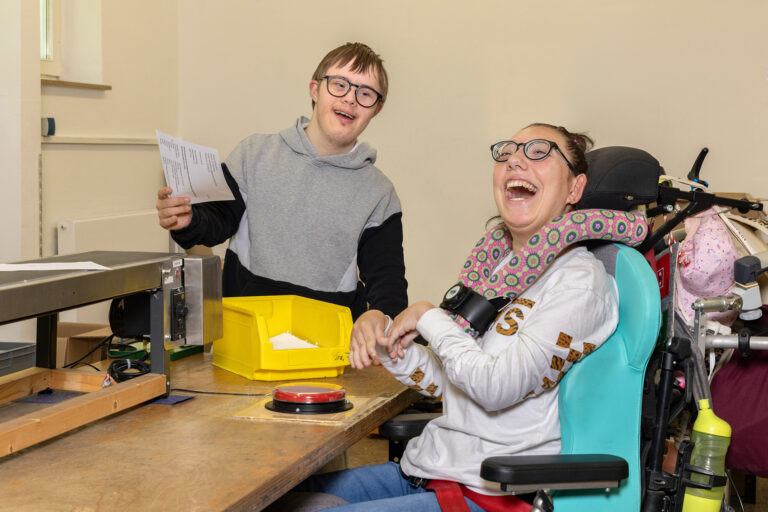 Zwei Teilnehmer lachen in die Kamera. Die Teilnehmerin sitzt im Rollstuhl und kann mit einem Knopf ein Förderband aktivieren. Der Teilnehmer legt gerade Papier auf das Förderband.
