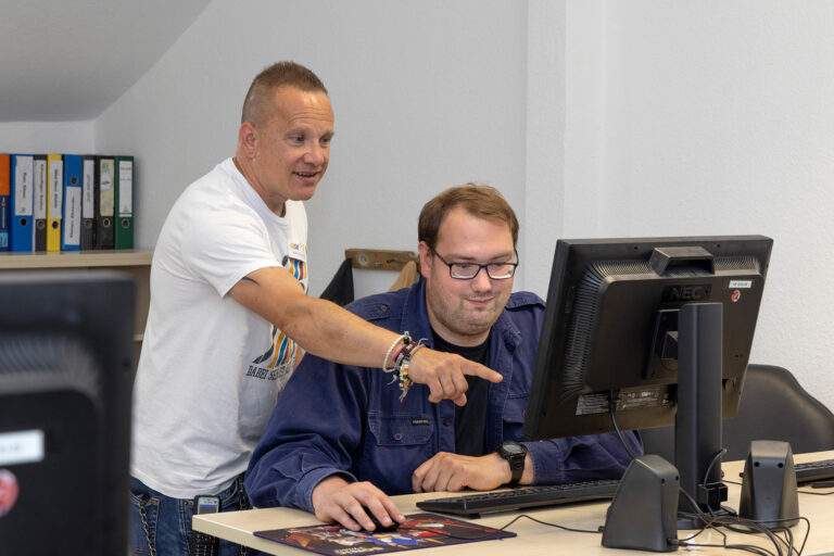 Ein Teilnehmer sitzt vor einem Computermonitor und hat eine Computermaus in der Hand. Ein Gruppenleiter steht hinter ihm und zeigt auf etwas auf dem Monitor.
