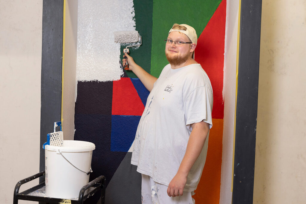 Ein Teilnehmer der Malergruppe streicht mit einer Farbrolle weiße Farbe über eine farbige Fläche und lächelt freundlich in die Kamera.