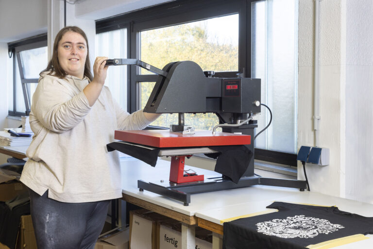 In der Druckerei bedient eine Werkstattmitarbeiterin die Maschine für den Druck auf Textilien. Daneben liegt ein bereits fertig gedrucktes T-Shirt.
