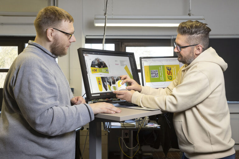 In der Druckerei bespricht ein Gruppenleiter mit einem Werkstattmitarbeiter die weiteren Schritte bei der Bedruckung einer Tasse.