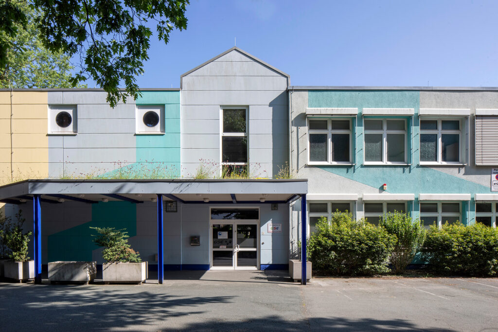 Der Eingangsbereich der GSE-Werkstatt Dellwig ist zu sehen. Ein hellgrau-türkis gestrichenes Gebäude mit einem Vordach ist zu sehen.