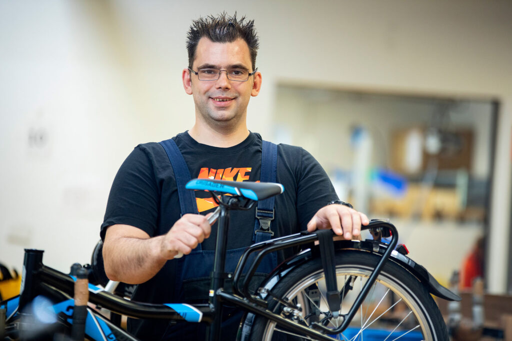 Ein Werkstattmitarbeiter schraubt einen Sattel an ein Kinderfahrrad und lächelt freundlich in die Kamera.