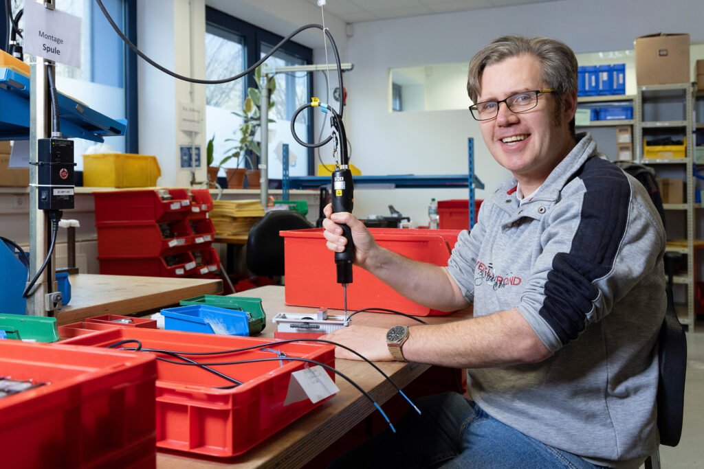 Ein Werkstattmitarbeiter montiert etwas an einem Arbeitsplatz. Er blickt in die Kamera und lächelt.