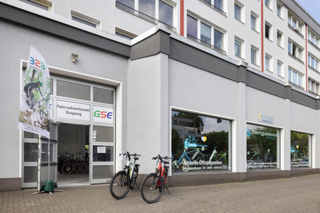 Die GSE-Fahrradwerkstatt Rüttenscheid ist in einem grauen Gebäude mit großen Schaufenstern. Es stehen zwei Fahrräder vor dem Eingang der Fahrradwerkstatt.